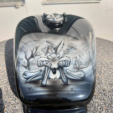 Ein schwarzer Motorrad-Kraftstofftank mit einem gezeichneten Cartoon-Charakter, wahrscheinlich ein Kojote, in einer Wüstenumgebung.