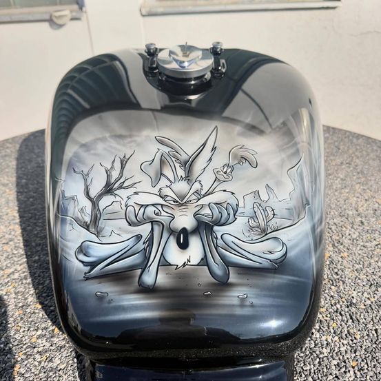 Ein schwarzer Motorrad-Kraftstofftank mit einem gezeichneten Cartoon-Charakter, wahrscheinlich ein Kojote, in einer Wüstenumgebung.