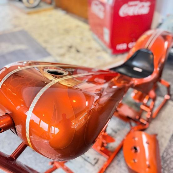 Eine Nahaufnahme eines orangefarbenen Motorrads mit glänzendem Finish, das den Kraftstofftank und Teile auf dem Boden zeigt. Im Hintergrund steht eine rote Coca-Cola-Box.