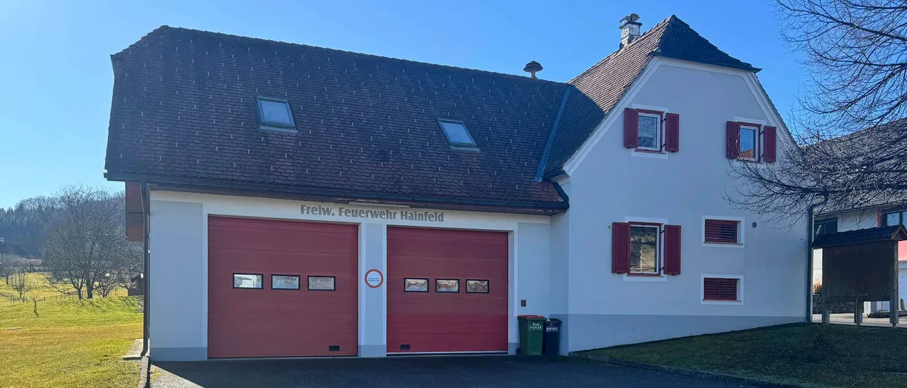 Ein weisses Feuerwehrhaus mit zwei roten Türen, jeweils mit kleinen Fenstern, und der Aufschrift Freiw. Feuerwehr Hainfeld an der Wand. Davor stehen Mülleimer.
