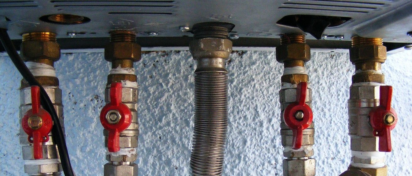 Nahaufnahme eines Warmwasserbereiters mit vier Rohren und roten Griffen, montiert an einer weißen Wand.