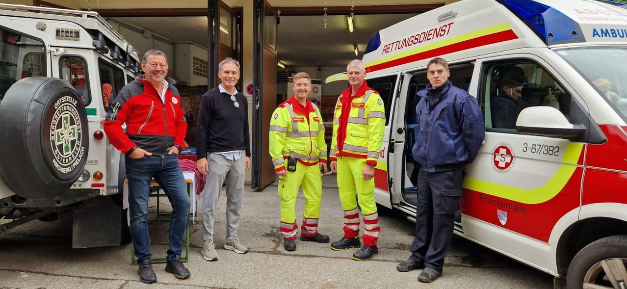 Fünf Männer stehen neben einem Feuerwehrauto. Die vier in Gelb tragen reflektierende Anzüge, während der Mann auf der linken Seite einen schwarzen Pullover und graue Hosen trägt.