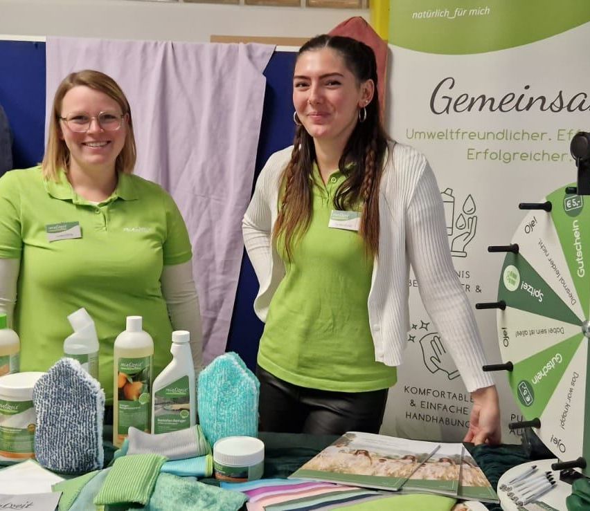 Zwei Frauen in grünen Shirts stehen hinter einem Tisch mit Reinigungsprodukten. Eine Frau lächelt und die andere schaut in die Kamera. Hinter ihnen ist ein Schild mit grünem Text.