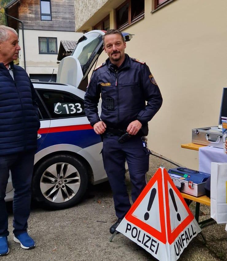 Ein Polizeibeamter steht neben einem Polizeiauto mit der Nummer 133, neben einem Mann in einem blauen Mantel. Dahinter befinden sich ein Polizeischild, eine Aktentasche und ein Tisch.