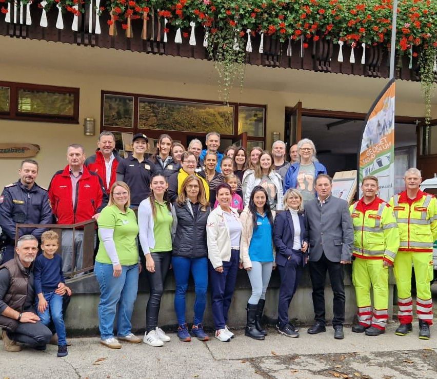 Eine Gruppe von Menschen steht vor einem Gebäude mit Blumen. Einige tragen Jacken, andere tragen Warnschutzanzüge. Ein Flaggen wird in der Nähe des Eingangs platziert.