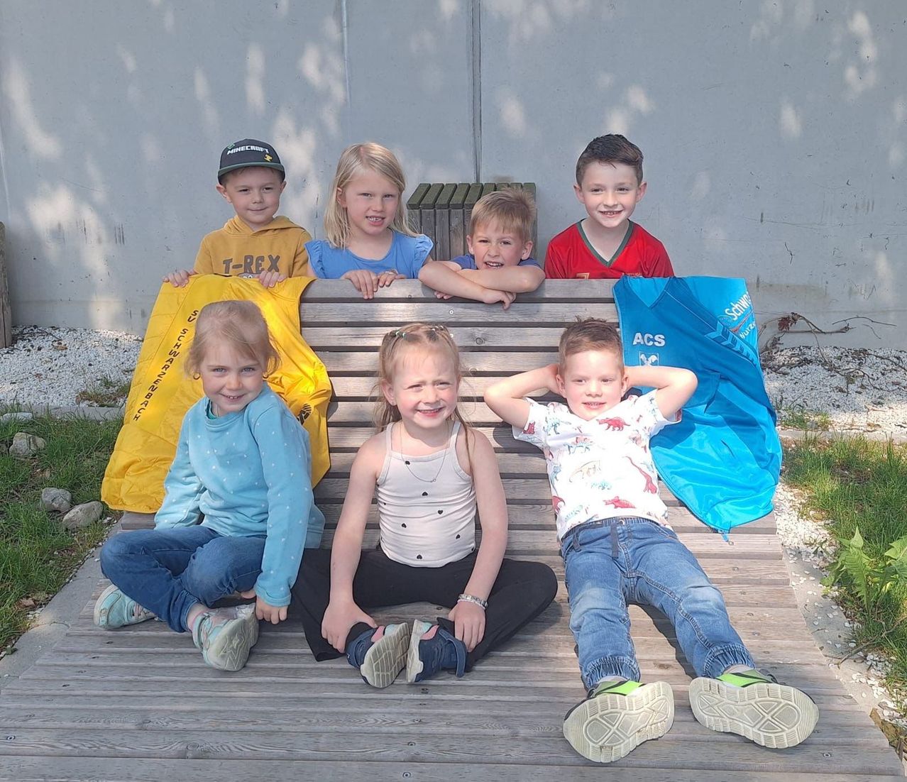 Sechs Kinder sitzen auf einer Holzbank und lächeln für ein Foto. Sie halten blaue und gelbe Taschen. Der Junge rechts trägt ein rotes Shirt, während die anderen Freizeitkleidung tragen.