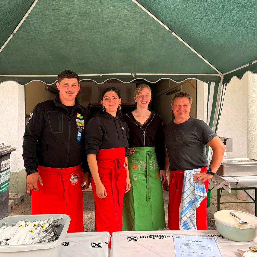 Vier Personen stehen unter einem Zelt, lächeln und posieren für ein Foto. Sie tragen Schürzen mit Logos und stehen neben einem Tisch mit Essen. Links steht ein Mülleimer.