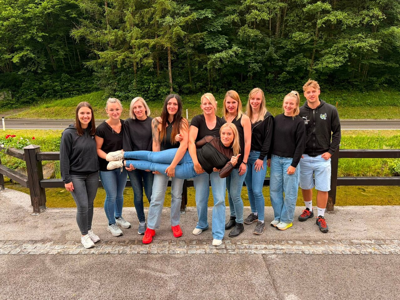 Eine Gruppe von Frauen und zwei Männern posiert für ein Foto. Eine Frau sitzt auf den Schultern einer anderen Frau. Sie tragen alle Schwarz und Jeans. Hinter ihnen steht ein Holzzaun.