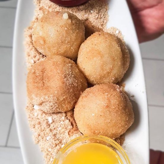 Ein weißer Teller mit vier goldbraunen, knusprigen frittierten Teigbällen, bestreut mit braunem Zucker, begleitet von einer kleinen Schüssel gelber Sauce.