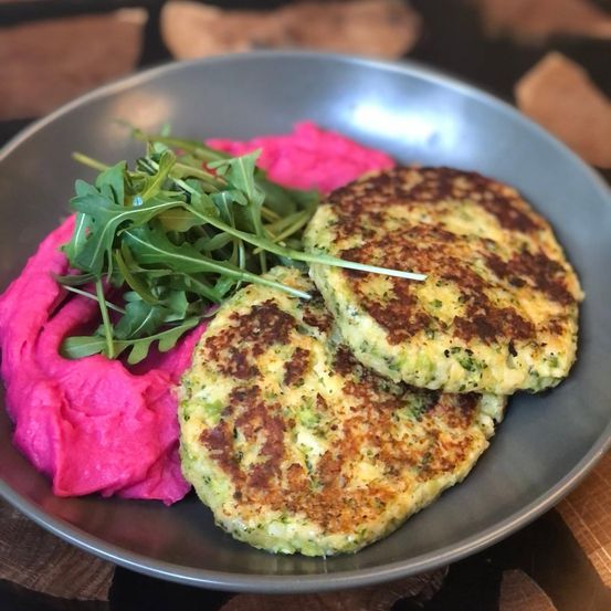 Ein Teller mit zwei goldbraunen Brokkoli-Krapfen, rosa Rübenpüree und Rucola auf einem grauen Teller.