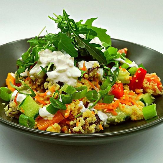 Eine Schüssel Couscous-Salat mit Rucola, Gurken, Karotten und roten Paprikaschoten, gekrönt von einem Klecks Joghurt.