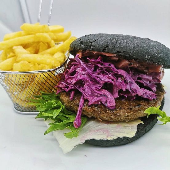 Ein schwarzer Burger mit rotem Kraut und Salat, serviert mit einem Korb goldener Pommes auf einem weißen Teller.