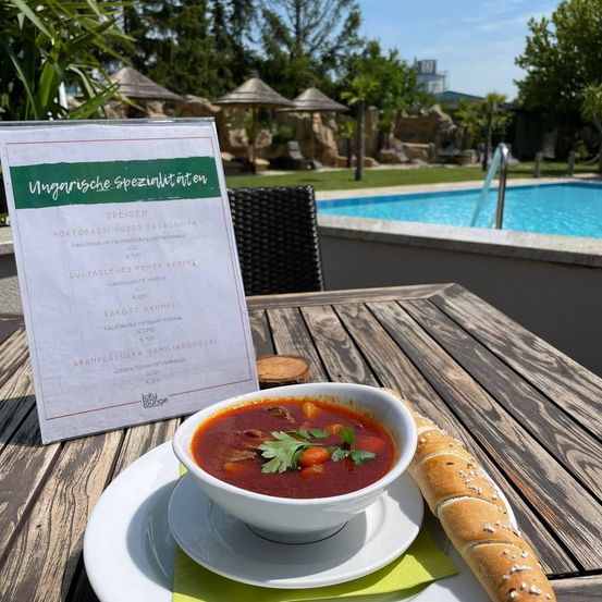 Eine Schüssel Suppe mit einem Stück Brot auf einem Teller, ein Menükarten auf dem Tisch und ein Schwimmbad im Hintergrund.