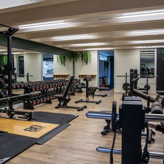 Ein gut beleuchteter Fitnessstudio-Innenraum mit verschiedenen Trainingsgeräten, einschließlich Hanteln, Bänken und einer Langhantel. Das Fitnessstudio hat Holzböden und weiße Wände.
