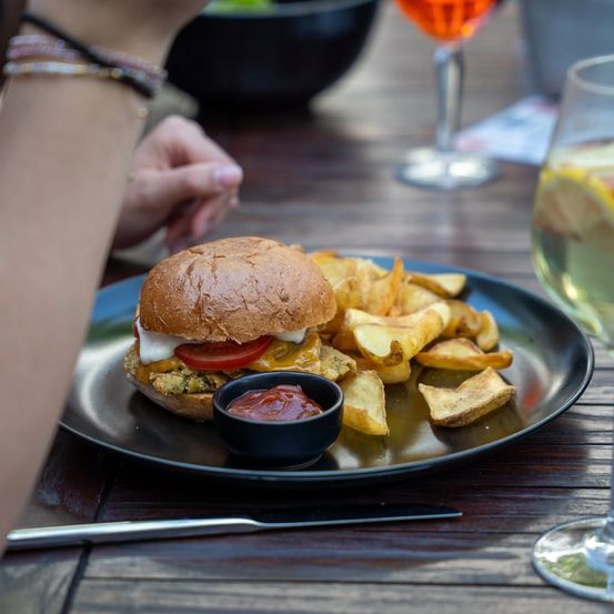 Ein Nahaufnahme eines Tellers mit einem Veggie-Burger, Pommes und einer kleinen Schüssel Ketchup auf einem Holztisch, mit einem Glas Wein und einem Weinglas im Hintergrund.