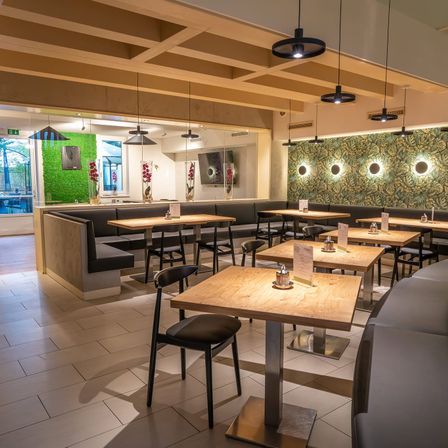 Ein Innenrestaurant mit Holztischen, schwarzen Stühlen und einer grünen Wand mit tropischem Blatttapeten. Der Raum hat ein modernes Design mit hängenden Lampen und einem gefliesten Boden.