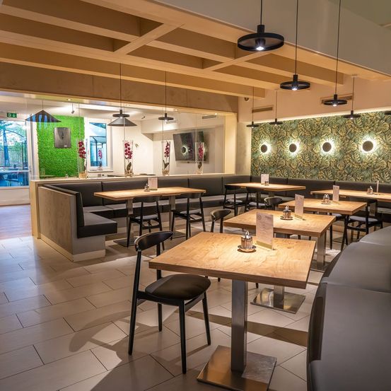 Ein Innenrestaurant mit Holztischen, schwarzen Stühlen und einer grünen Wand mit tropischem Blatttapeten. Der Raum hat ein modernes Design mit hängenden Lampen und einem gefliesten Boden.
