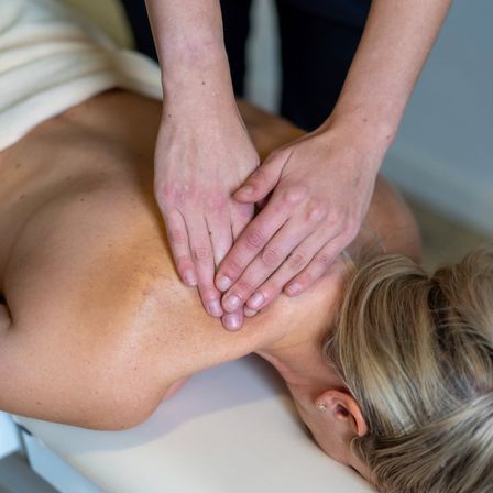 Eine Frau liegt auf einem Massagetisch und bekommt von einer Person in schwarzer Kleidung eine Nacken- und Schultermassage.