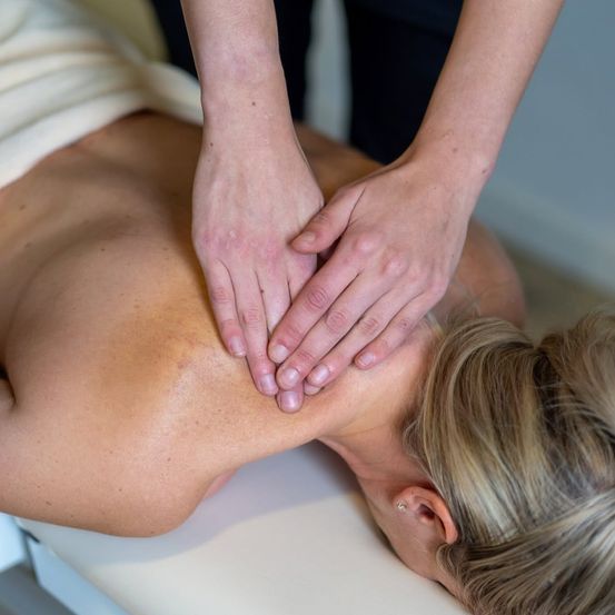 Eine Frau liegt auf einem Massagetisch und bekommt von einer Person in schwarzer Kleidung eine Nacken- und Schultermassage.