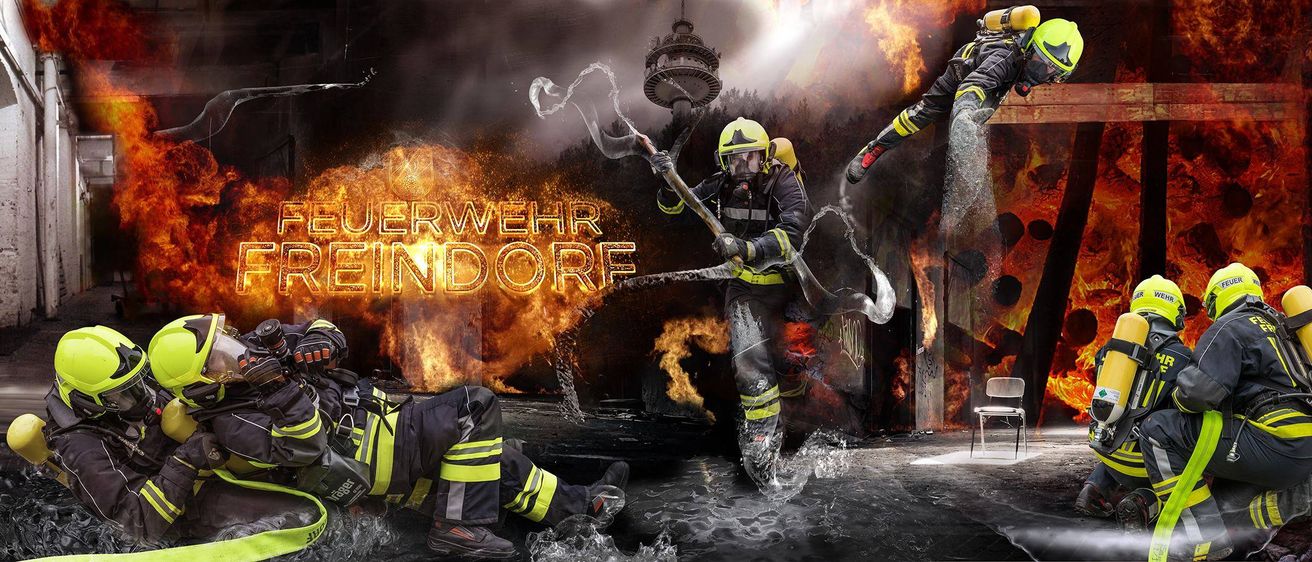Ein Feuerwehrteam im Einsatz, ein Feuerwehrmann sprüht Wasser, während ein anderer auf dem Boden liegt, beide vor einem brennenden Hintergrund.