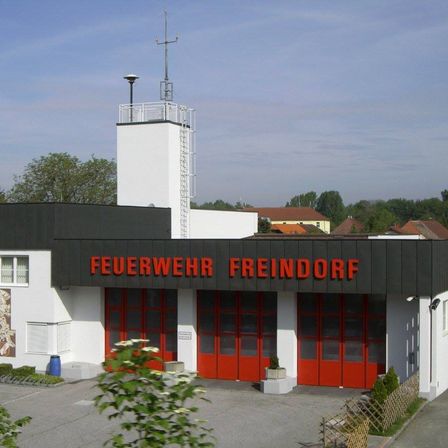 Ein Feuerwehrhaus in Freindorf mit einem Turm und einer Wand mit einem Wandbild. Es hat rote Türen und ein Schild mit der Aufschrift 'FEUERWEHR FREINDORF'.
