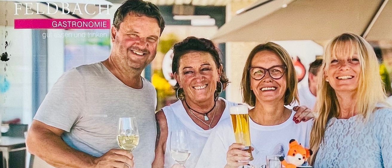 Plakat für eine Sommerveranstaltung in Feldbach, Österreich, mit vier lächelnden Erwachsenen, die Getränke und eine Tablett mit einem Plüschtier halten. Das Plakat enthält den Namen, das Datum und die Website der Veranstaltung.