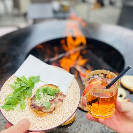 Eine Person hält ein Glas mit einem Getränk und einem Strohhalm. Eine andere hält einen Teller mit einem Sandwich. Im Hintergrund brennt ein Feuer.