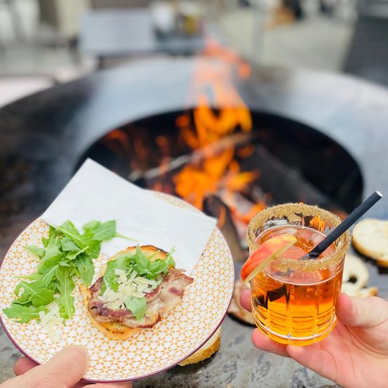 Eine Person hält ein Glas mit einem Getränk und einem Strohhalm. Eine andere hält einen Teller mit einem Sandwich. Im Hintergrund brennt ein Feuer.