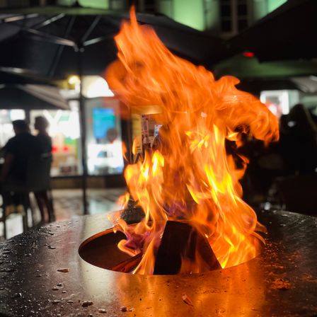 Ein Feuer brennt in einem äußeren Kamin bei Nacht, mit Flammen, die nach oben springen. In der Nähe sitzen Leute an Tischen unter Schirmen.