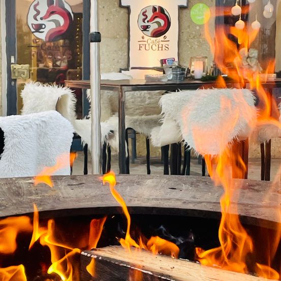 Ein gemütliches Café mit einem Kamin, umgeben von Stühlen mit Fellkissen. In der Mitte brennt ein Feuer hell.
