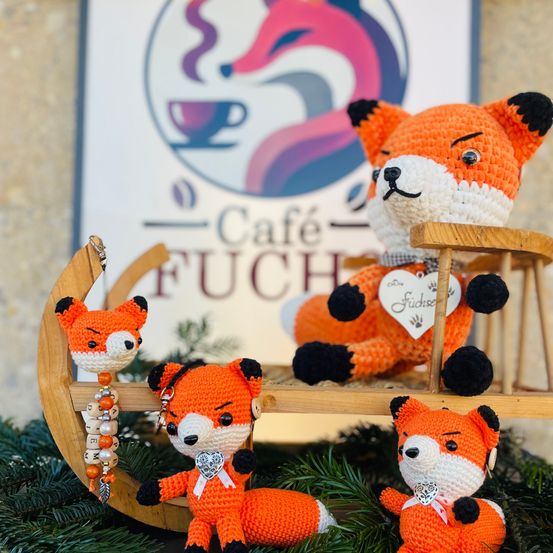Drei gehäkelte orange Füchse sitzen auf einem hölzernen Schlitten vor einem Schild, das 'Cafe Fuchs' steht. Jeder Fuchs trägt eine Schleife, und der mittlere hat einen herzförmigen Anhänger.