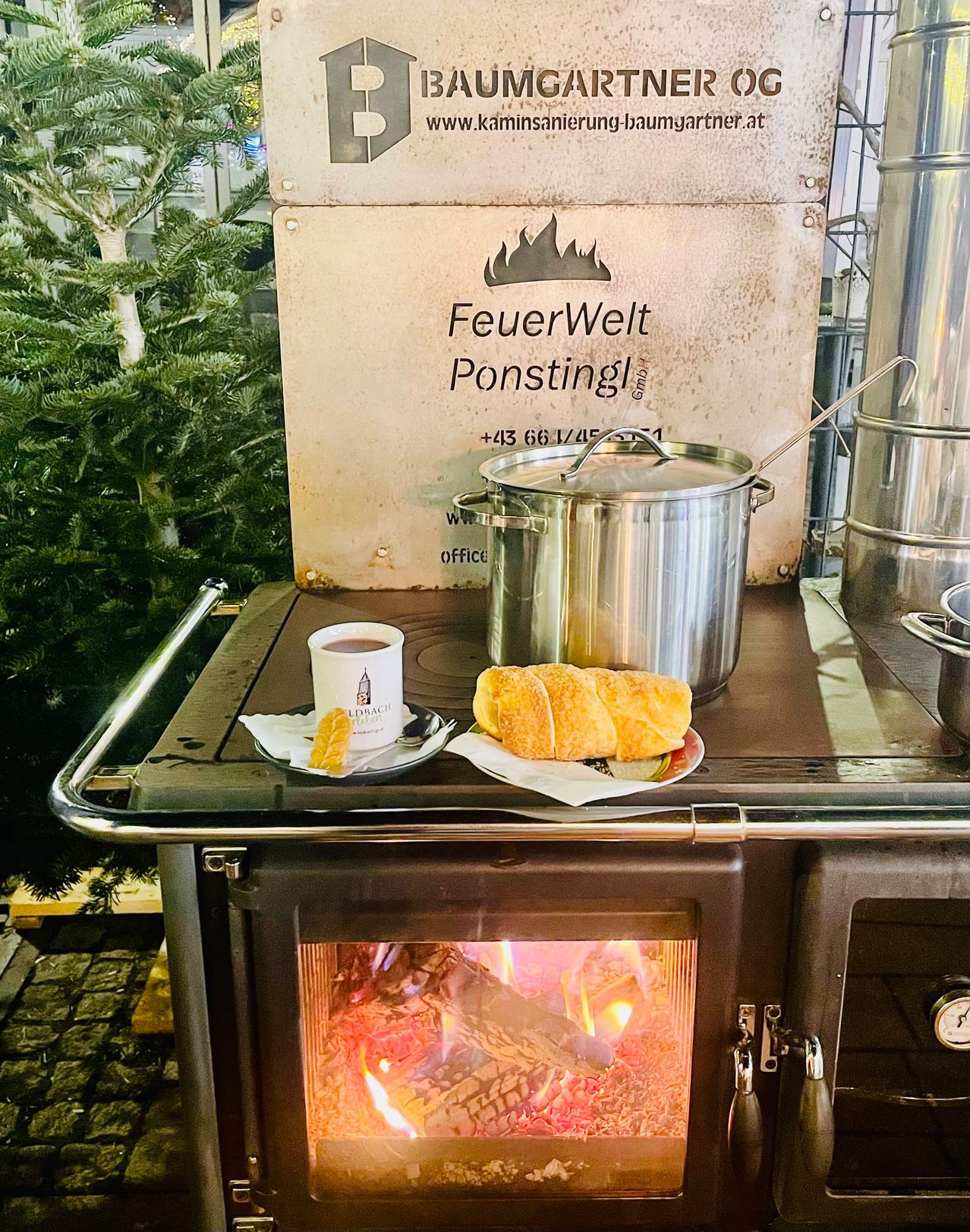 Ein Kamin mit einem Topf darauf, neben einer Kaffeetasse und einem Teller mit Croissants. Dahinter befindet sich ein Schild für FeuerWelt Ponstingl.