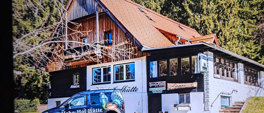 Ein blauer Van mit einem Truthahn-Design ist vor einem rustikalen Haus mit einem braunen Dach und weißen Wänden geparkt. Das Haus hat mehrere Fenster und ein hölzerne Schild, das 'Hütte' steht.