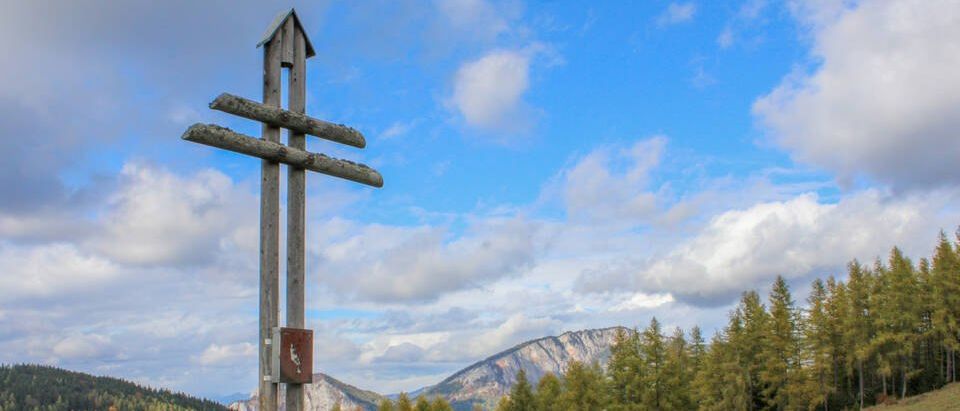 Ein Holzkreuz steht auf einem grasbewachsenen Hügel mit Blick auf Berge und einen blauen Himmel mit Wolken.