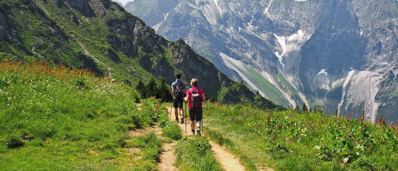 Zwei Personen wandern auf einem Pfad, der von Bergen umgeben ist. Sie tragen Rucksäcke und halten Trekkingstöcke. Der Pfad ist schmal und führt den Berg hinauf.