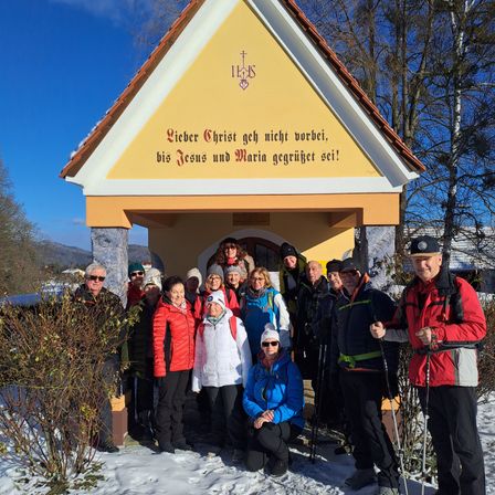 Eine Gruppe von Menschen, für den Winter gekleidet, steht vor einer Kapelle und lächelt für ein Foto. Die Kapelle hat eine gelbe Wand mit einem Kreuz und einer deutschen Inschrift.
