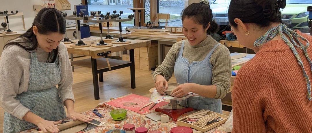 Drei Frauen arbeiten gemeinsam an einem Tisch, jede mit einem Stück Ton, umgeben von Bastelmaterialien. Sie tragen Schürzen und scheinen in einem Töpferkurs engagiert zu sein.