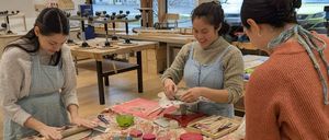 Drei Frauen arbeiten gemeinsam an einem Tisch, jede mit einem Stück Ton, umgeben von Bastelmaterialien. Sie tragen Schürzen und scheinen in einem Töpferkurs engagiert zu sein.
