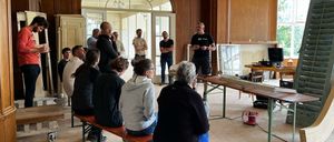 Eine Gruppe von Menschen ist in einem Raum versammelt, wahrscheinlich für eine Besprechung. Ein Mann steht und spricht, möglicherweise eine Präsentation haltend. Der Raum hat Holzbänke, Schreibtische und Schränke. Es hängen auch Lampen von der Decke.