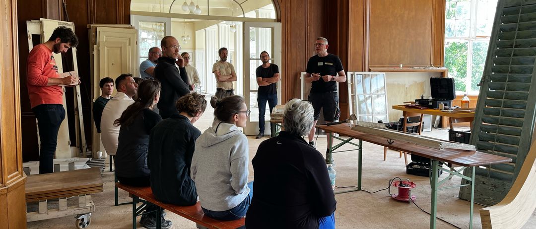 Eine Gruppe von Menschen ist in einem Raum versammelt, wahrscheinlich für eine Besprechung. Ein Mann steht und spricht, möglicherweise eine Präsentation haltend. Der Raum hat Holzbänke, Schreibtische und Schränke. Es hängen auch Lampen von der Decke.