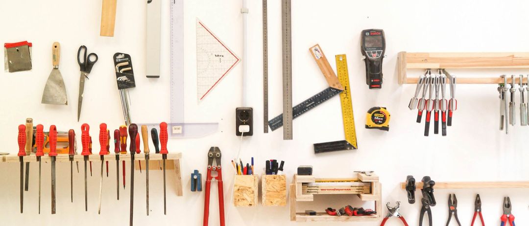 Eine weiße Wand zeigt verschiedene Werkzeuge und Geräte, darunter Messwerkzeuge, ein Wasserwaage, eine Säge und einen Hammer, alle ordentlich auf Holzregalen organisiert.