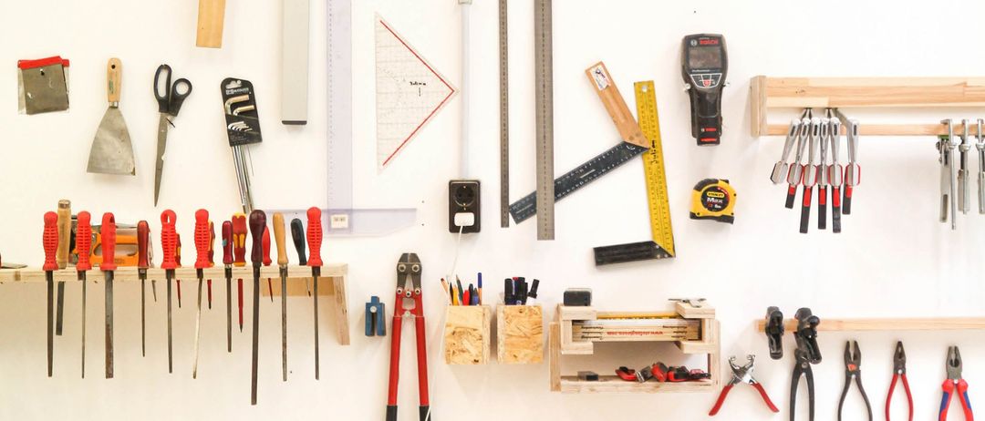 Eine Vielzahl von Werkzeugen und Messinstrumenten sind an einer weißen Wand organisiert, einschließlich Hämmer, Schraubendreher und eine Wasserwaage. Andere Werkzeuge wie Zangen, Drahtschneider und ein Maßband sind ebenfalls vorhanden.
