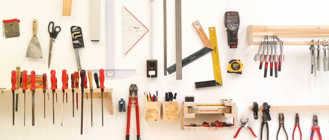 Eine weiße Wand mit verschiedenen Werkzeugen, darunter ein Satz Schraubendreher, Zangen, Messbänder und andere Instrumente. Hölzerne Regale halten die Werkzeuge und die Wand ist weiß gestrichen.