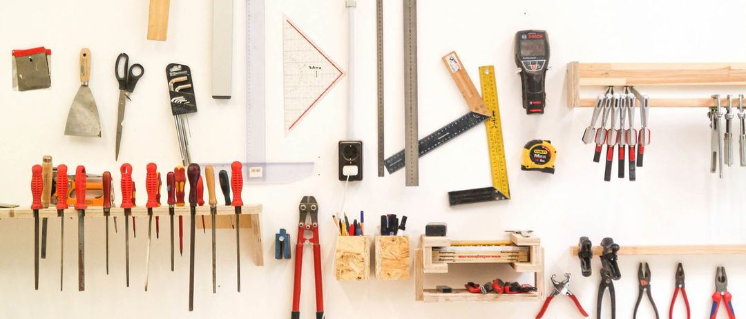 An einer weißen Wand ist eine Auswahl an Werkzeugen zu sehen, darunter Schraubendreher, Zangen, Maßbänder und verschiedene andere Geräte. Holzregale und Haken halten die Werkzeuge ordentlich.
