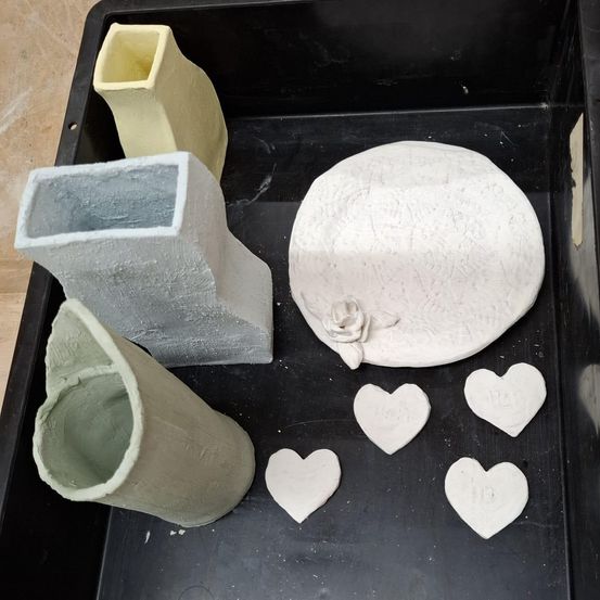 Ein schwarzer Tablett hält verschiedene Tonskulpturen, darunter eine große weiße Keramikschale, eine hohe gelbe Vase, ein graues zylindrisches Stück, ein grünes zylindrisches Stück und mehrere herzförmige Tonstücke. Das Tablett steht auf einer Holzoberfläche.