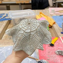 Eine Hand hält ein Keramikstück mit einem komplexen Design in einer Werkstatt. Verschiedene Werkzeuge und Materialien sind auf den Holztischen verstreut. Dahinter ein Fenster mit Jalousien und ein Blick aufs Freie.