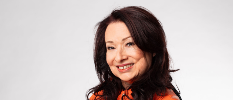 Eine Frau in einer orangefarbenen Lederjacke lächelt fröhlich, ihre Hände vor sich verschränkt. Sie hat dunkelbraunes Haar, das in lockeren Wellen gestylt ist, und ihr Outfit vermittelt einen lässigen, aber stilvollen Eindruck.