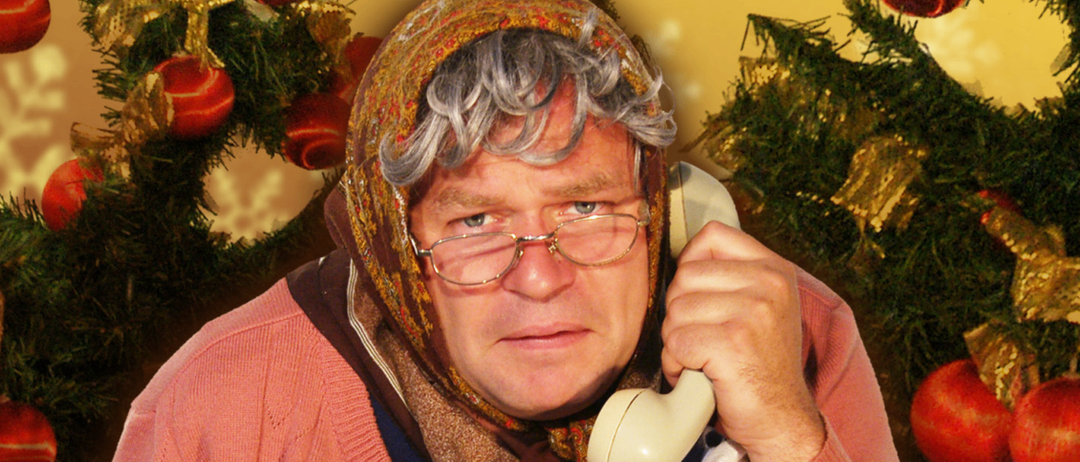 Eine ältere Person in einem rosa Pullover und Schal spricht am weißen Telefon. Hinter ihm sind Weihnachtsdekorationen sichtbar.