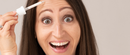 A woman with brown hair is smiling and holding a bottle of liquid in her hand. She has a white pipette in her right hand on her forehead.