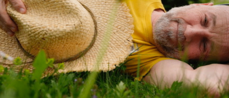 A man lies on the grass with his hand on a straw hat. He is wearing a yellow shirt and has a white beard. The grass is green and lush.